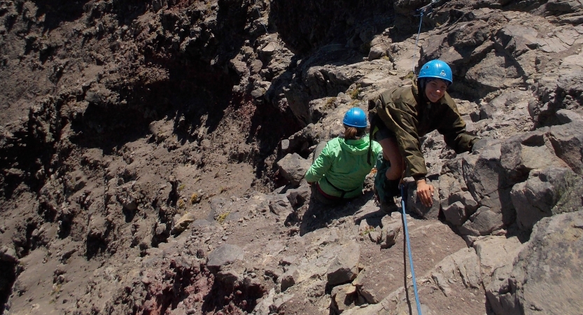 A person wearing rock climbing gear looks up at the camera and smiles while rock climbing. Below them, another person faces away from the camera.